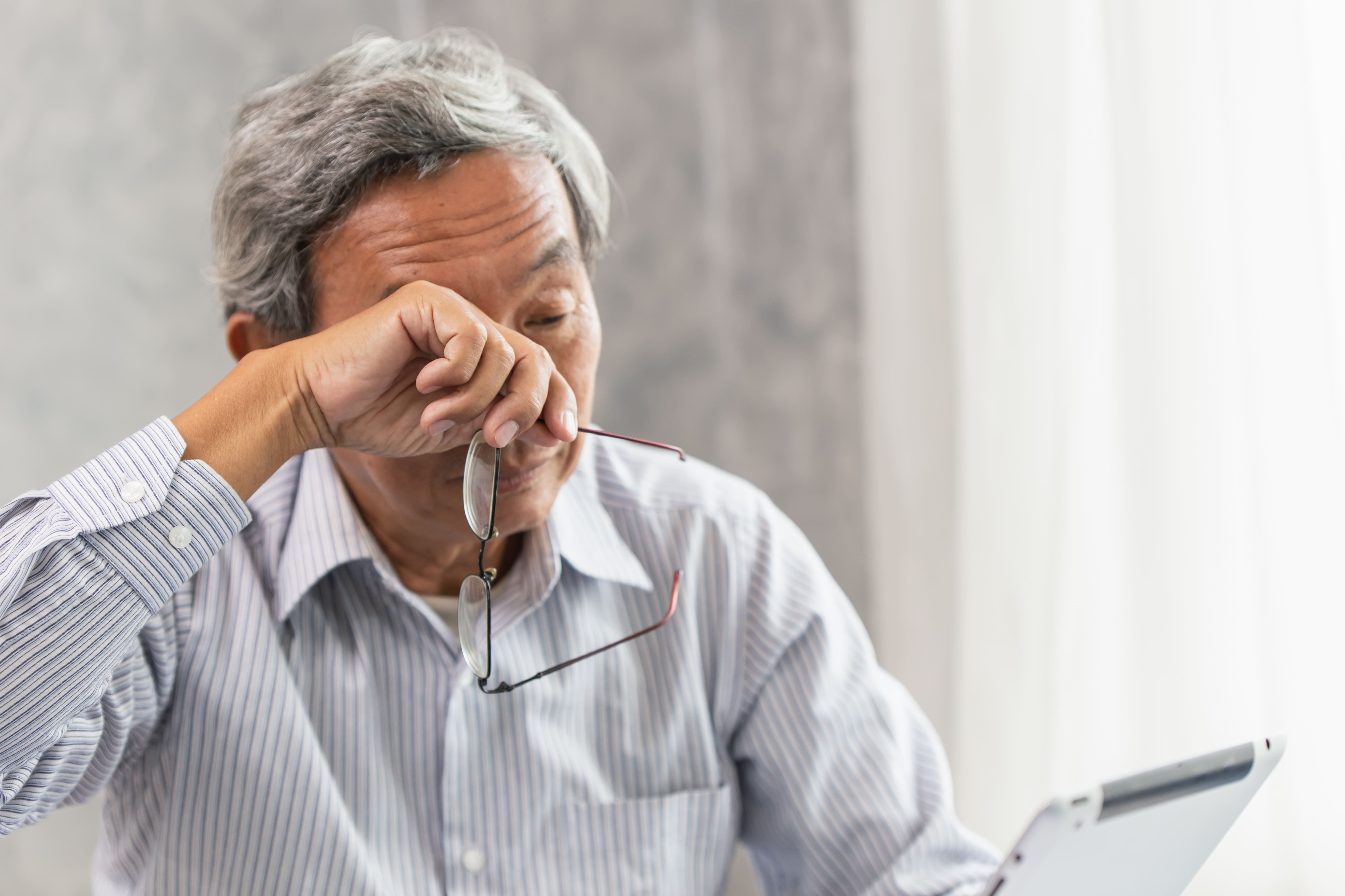 Elderly man rubbing tired eyes while using a tablet, highlighting signs that may require cataract treatment in Singapore.