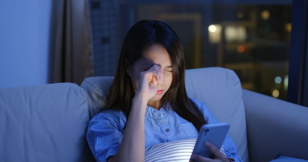 Woman rubbing an irritated eye while using her phone at night, highlighting symptoms that may require a dry eyes specialist in Singapore.